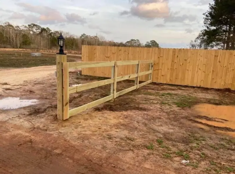 A three-rail pine fence meeting a tall pine privacy fence at a corner post with a lamp, on a freshly cleared lot