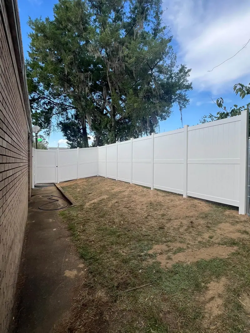 A long run of white vinyl privacy fence along a brick house under a big shade tree, installed by Southern Line Fence & Finish